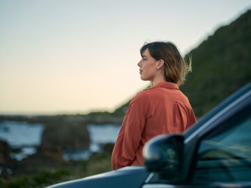 A woman stands beside her car and looks out at the water. She is calm