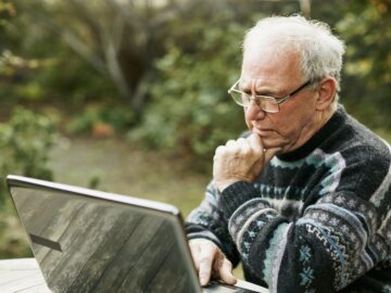 An older man with white hair is sat before his laptop, outside, showing distrust at what he sees.