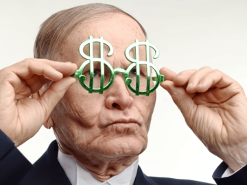 An older man holding dollar sign glasses up to his eyes.