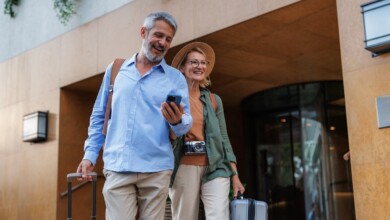 A happy couple rolls their luggage to a travel destination as the man references his phone.